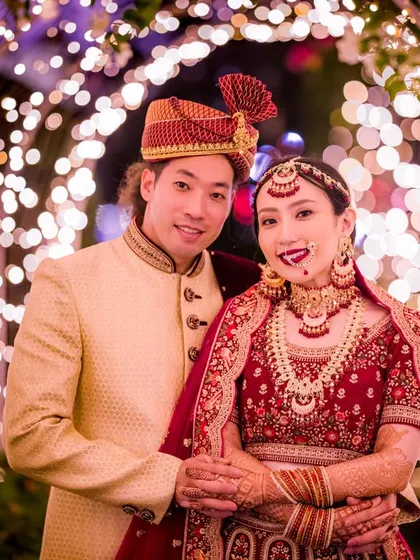 A classic portrait of a couple in stunning traditional wedding attire, framed by a beautiful arch of fairy lights. The warm glow highlights the intricate details of their outfits.