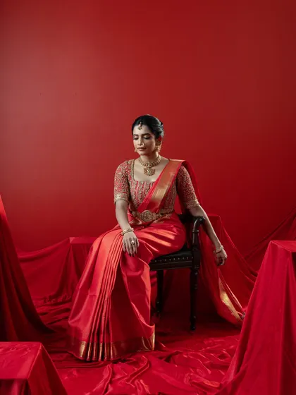 Seated with regal poise, the bride showcases the beautiful drape of the red Kanjeevaram saree. The rich fabric pools around her, creating a scene of classic elegance and cultural richness.