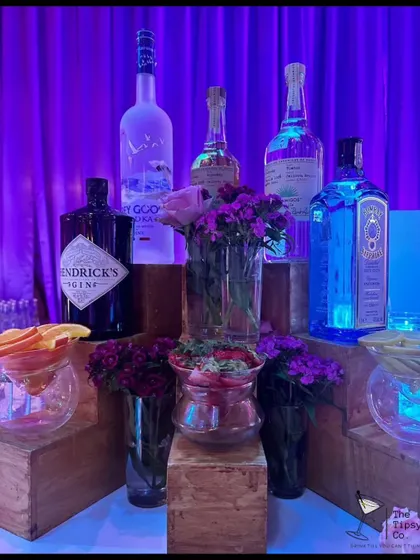 A beautiful display of premium gins and vodkas, accented with fresh purple flowers. The wooden block risers and soft lighting create a warm and inviting gin station.