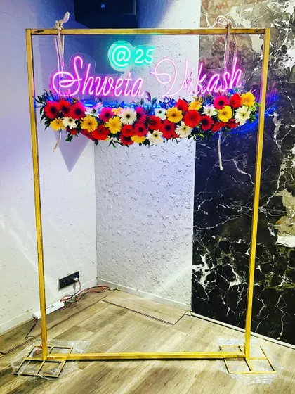 A personalized and colorful photo booth frame. A custom neon sign with the names 'Shweta Vikash' is mounted on a golden rectangular frame and decorated with a line of vibrant, multi-colored flowers.