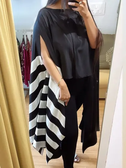 A client trying on our "Monochrome Mania" cape. This striking piece features a solid black top with dramatic, flowing side panels in a bold black and white stripe pattern, made from a cotton and satin blend.
