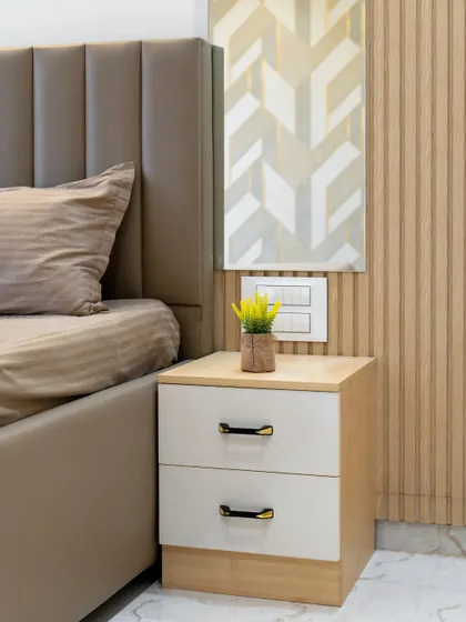 A close-up of a bedside table in a neutral-themed bedroom. The design combines a light wood finish with a cream-colored drawer, and the wall features a mix of textures.
