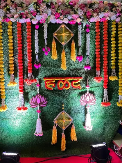 A detailed view of a Haldi backdrop against an artificial green wall. The decor features hanging garlands of red and yellow flowers, traditional lotus-themed hangings, and a prominent 'हळद' sign in the center.