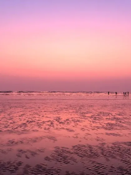 A beautiful photograph of a Mumbai beach at sunset, with the wet sand reflecting the soft pink and purple hues of the sky.