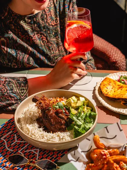 A lifestyle shot at Lyla Bombay, focusing on a bowl of food and a bright red cocktail. The colorful patterns in the decor and clothing add to the lively atmosphere.