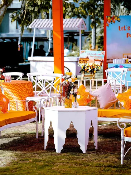 A relaxed outdoor seating arrangement with white wrought-iron furniture and bright orange cushions, perfect for a sunny Haldi celebration.