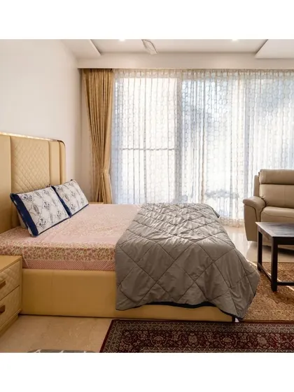 A serene bedroom where sunlight streams through sheer curtains. The quilted leatherette bed with a gold-trimmed headboard adds a touch of luxury, while the comfortable recliner creates a perfect corner for morning coffee.