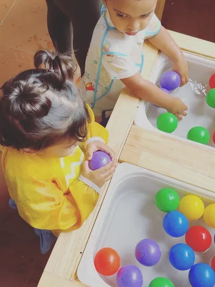 A child holds a purple ball from the water table, examining it closely. This simple action is part of the sensory exploration process, helping them understand concepts like shape, color, and texture.