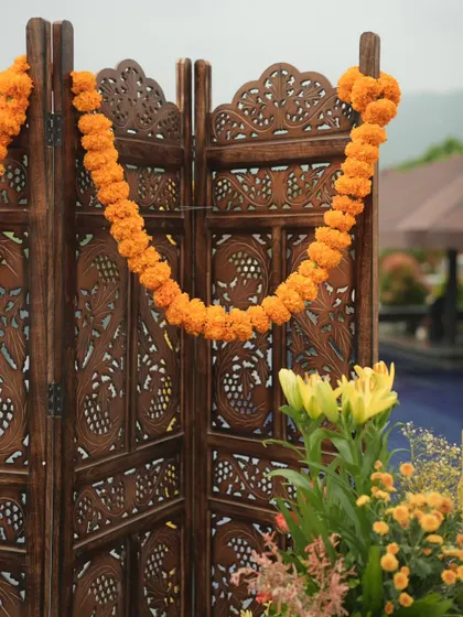 A close-up of the intricately carved wooden screen used as a backdrop element, adorned with a simple marigold garland to enhance its traditional appeal.