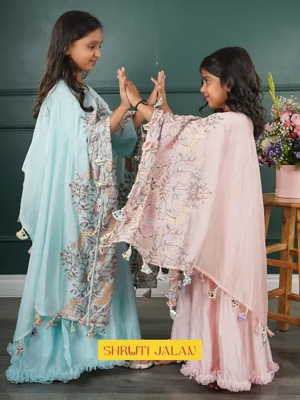 This image captures two sisters in my flowing cape sets, one in sky blue and the other in blush pink. The capes are adorned with printed floral motifs and delicate tassels, adding a whimsical touch to the outfits.