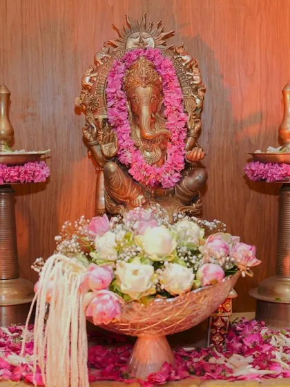 A close up of a Ganpati idol adorned with a fresh pink flower garland. The setup includes traditional brass lamps (samai) and a bowl of lotuses, creating a simple yet beautiful space for worship.