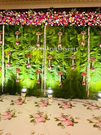 A classic South Indian backdrop made from fresh banana leaves. The stage is decorated with hanging floral tassels and a top border of colorful flowers, perfect for any auspicious event.