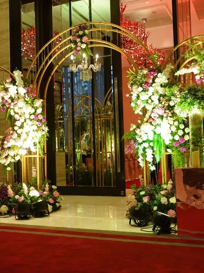Golden arches and a red carpet create a regal path for guests. This entrance is adorned with lush arrangements of pink, purple, and white flowers, offering a grand and colorful welcome to the wedding reception.