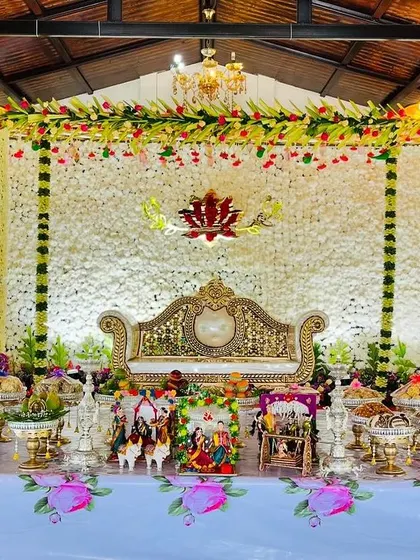 A pristine white floral backdrop provides an elegant setting for this Seemantham ceremony. The stage is adorned with a lotus cutout, traditional dolls, and a full arrangement of offerings.