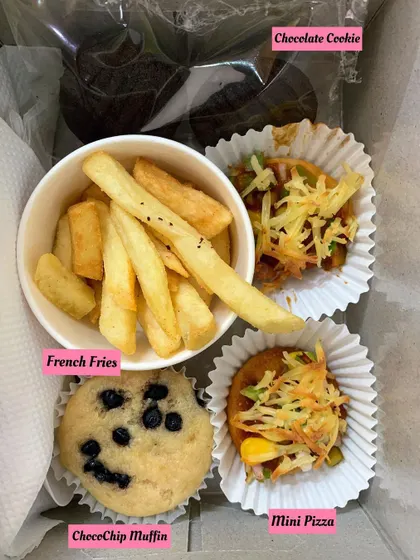 A close-up of a kid's party box with french fries, a choco-chip muffin, mini pizza, and a chocolate cookie.