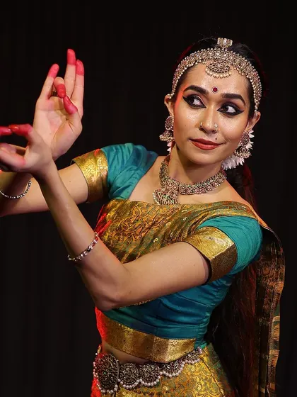 A close-up studio portrait capturing the intricate details of the performance look, from the temple jewelry to the expressive eyes and hand mudras.