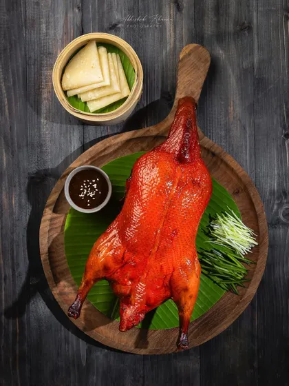 A whole roasted duck, styled on a wooden board with a banana leaf. The dark, moody lighting creates a dramatic and high-end feel for this signature dish.