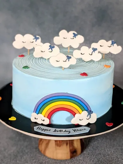 A dreamy chocolate cake with a "Twinkle Twinkle Little Star" theme. The light blue whipped cream frosting is decorated with a fondant rainbow and cute, sleepy cloud toppers.