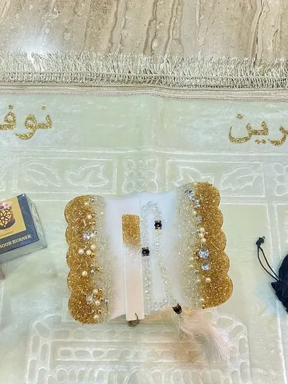 A personalized white and gold Quran holder set, including a bookmark and crystal tasbih, placed on a prayer mat embroidered with the clients' names.