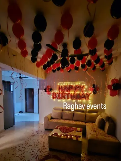 A cozy and romantic birthday decoration in a living room. The setup features red and black balloons on the ceiling, a 'Happy Birthday' foil balloon banner, and rose petals on the floor.