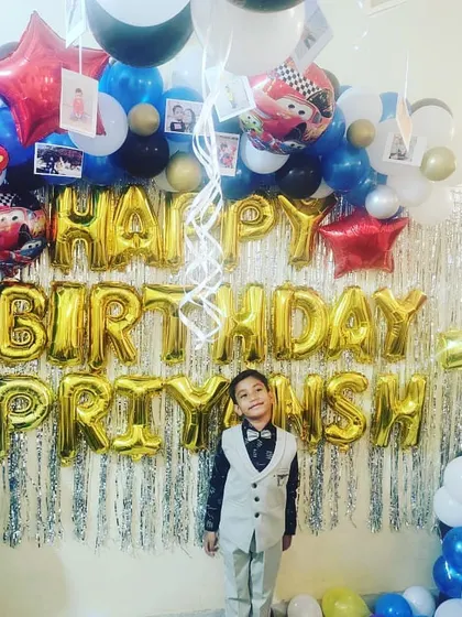 A 'Cars' themed 7th birthday party. The birthday boy stands proudly in front of a silver fringe backdrop, framed by a balloon arch with character cutouts and photos.