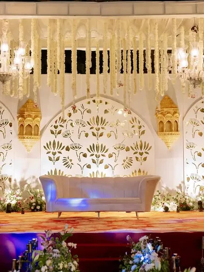 An elegant white and gold stage for a traditional ceremony, featuring intricate jaali work, hanging floral strings, and ornate lanterns.