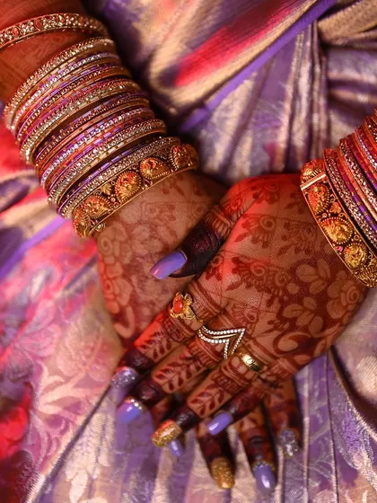 A close-up of the bride's hands, showcasing the rich mehendi stain against her purple saree and bangles.