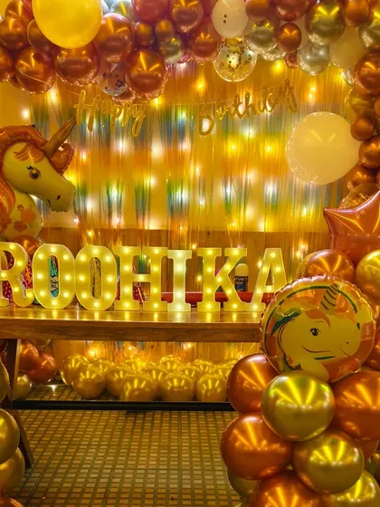 A glamorous unicorn-themed first birthday for Roohika. The decor features shiny chrome balloons in gold and bronze, a unicorn foil balloon, and her name in bright marquee letters.