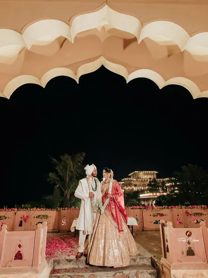 A wide angle view of the couple standing under a traditional archway, with the majestic palace in the distance. This shot establishes the scale and beauty of a destination palace wedding.