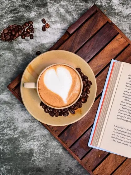 A classic coffee and book flat lay. The latte art, roasted beans, and open book create a narrative of a quiet, relaxing moment in a rustic cafe.