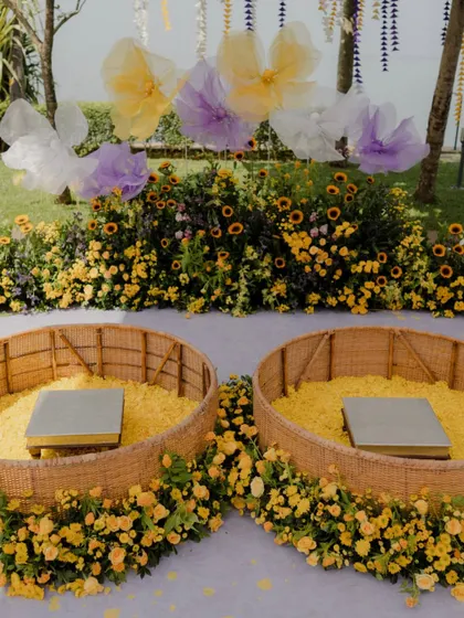 The twin seating arrangement for the couple at the pastel Haldi ceremony. The wicker baskets are filled with marigold petals and surrounded by a bed of sunflowers and other yellow blooms.