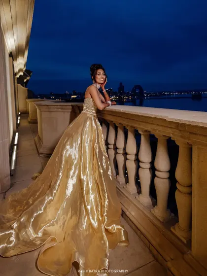 The bride in a shimmering golden gown looks out from a balcony at night, with the glittering Baku city skyline in the background.