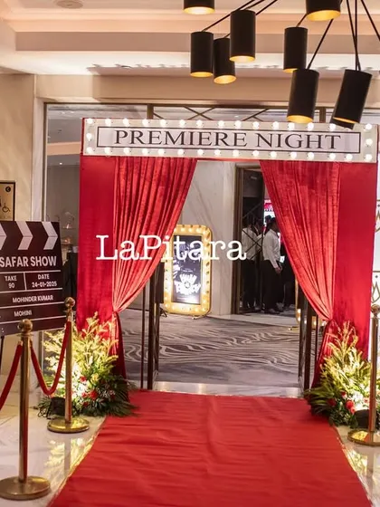 A red carpet 'Premiere Night' entrance for a Bollywood-themed 90th birthday party. The setup includes velvet ropes, a custom clapperboard, and movie poster displays.