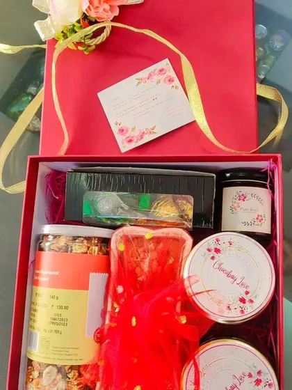 The inside of the red gift box, packed with an assortment of goodies. You can see jars of snacks, a box of chocolates, and other treats nestled in pink shredded paper.