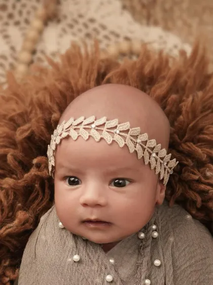 An expressive close-up of a wide-eyed newborn baby girl, wearing a delicate leaf-patterned headband on a soft, furry brown blanket.