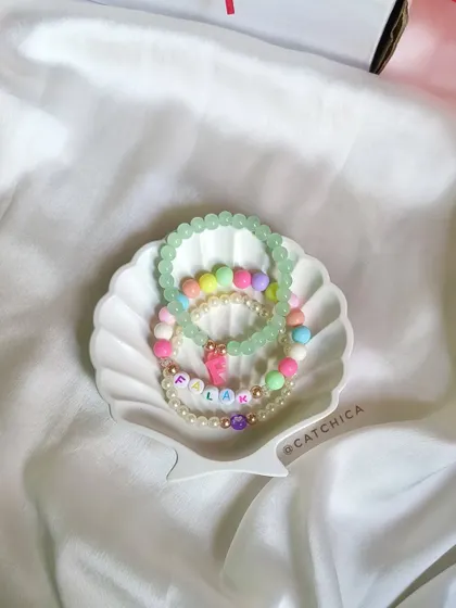 A stack of three personalized bracelets for Palak, displayed in a delicate shell dish. We love mixing pastel beads, pearls, and initial charms.