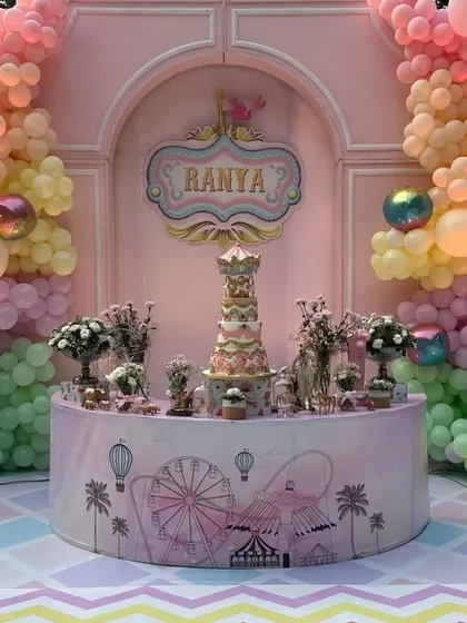 The central cake table, set against a pastel pink backdrop and surrounded by balloon garlands, with a magnificent carnival-themed cake.