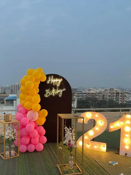 This is a duplicate of ID 21, 67, 91, 108, 121, 168, 206, 232, 239. A vibrant 21st birthday decoration on a rooftop. This setup features a balloon arrangement in hot pink and yellow, a 'Happy Birthday' neon sign, and large marquee numbers.