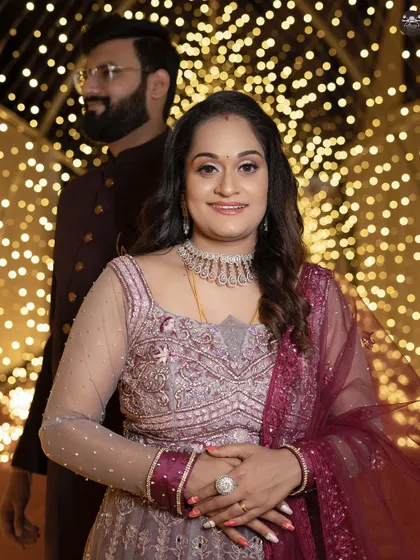 A beautiful portrait of a bride at her reception, smiling at the camera. The groom stands slightly behind her, out of focus, against a wall of golden bokeh lights, creating a lovely depth.
