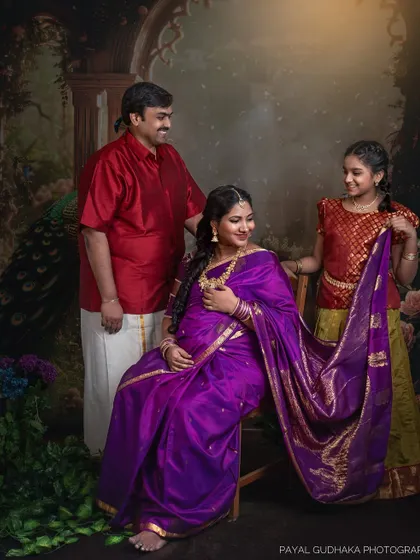A family portrait featuring an expecting mother in a purple saree, her husband, and their daughter. The daughter helps hold the saree's pallu, adding a sweet, interactive element to this traditional shoot.
