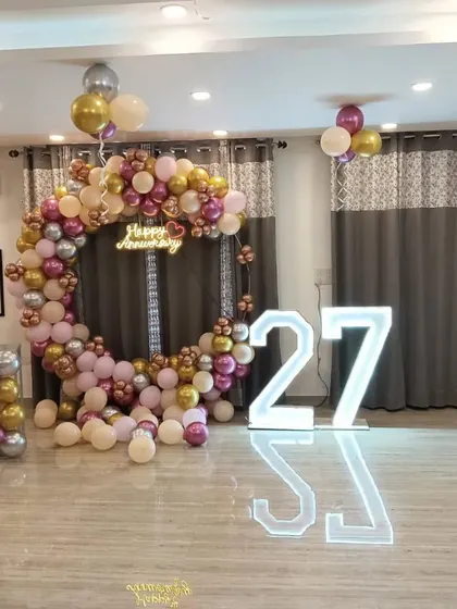 A 27th-anniversary celebration with a circular balloon arch in pink, gold, and silver. The setup includes large marquee numbers and floating balloons.