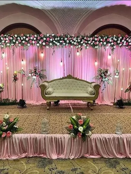 Here is a reception stage setup with a soft, romantic feel. I used a backdrop of pink drapes with hanging bulb lights, topped with a dense arrangement of pink and white flowers to create a warm and inviting space.