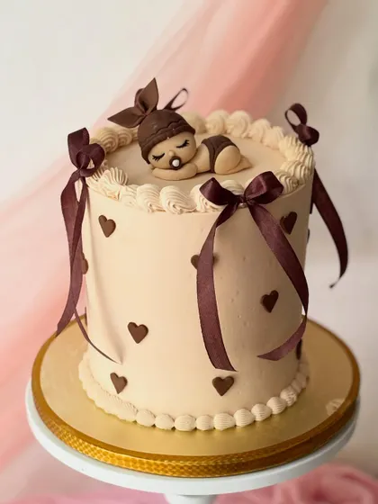 A cozy and cute baby shower cake in a warm brown color palette. The cake is decorated with hearts, ribbons, and a sleeping baby topper.