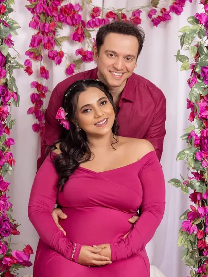 A vibrant and joyful studio maternity portrait. The happy couple is framed by a beautiful arch of bright pink bougainvillea flowers, with the mom-to-be glowing in a matching magenta dress.