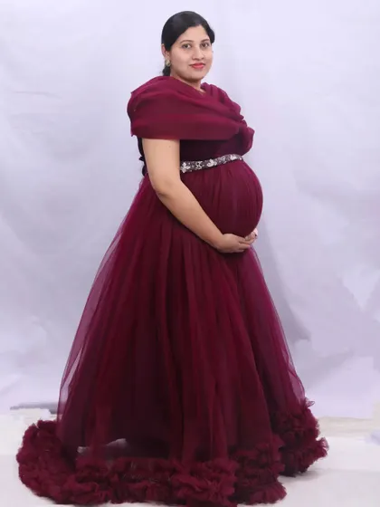 A classic portrait in a rich maroon gown with a ruffled train. The off-shoulder design and embellished belt add elegant details to this maternity look.