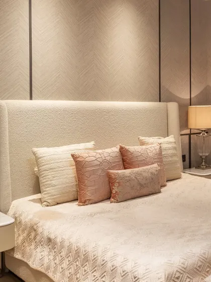 A close-up of a high-backed upholstered headboard in a soft, neutral fabric. The layered pillows in complementary pastel tones add a touch of color and texture, creating a plush and inviting feel.