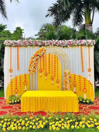A modern and stylish Haldi stage setup for an outdoor event. This design uses a combination of white drapes, geometric golden arches, and marigold flower arrangements for a fresh and contemporary look.