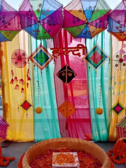 A vibrant Haldi ceremony space featuring a multi-colored backdrop with hanging kites and umbrellas. The 'urli' is filled with flower petals, ready for the rituals.