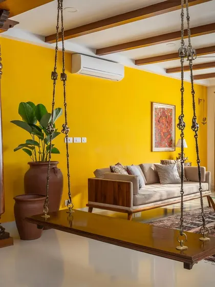 The traditional wooden swing and antique Chettinad pillar in 'Vasudhalayah' are the stars of the living room, set against a vibrant yellow wall that represents the bold Indian color palette.