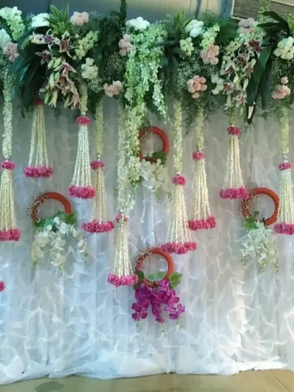 This is a traditional backdrop design often used for poojas or pre-wedding ceremonies. It features hanging strings of jasmine and mogra flowers (kaleeras) against a white ruffled curtain.